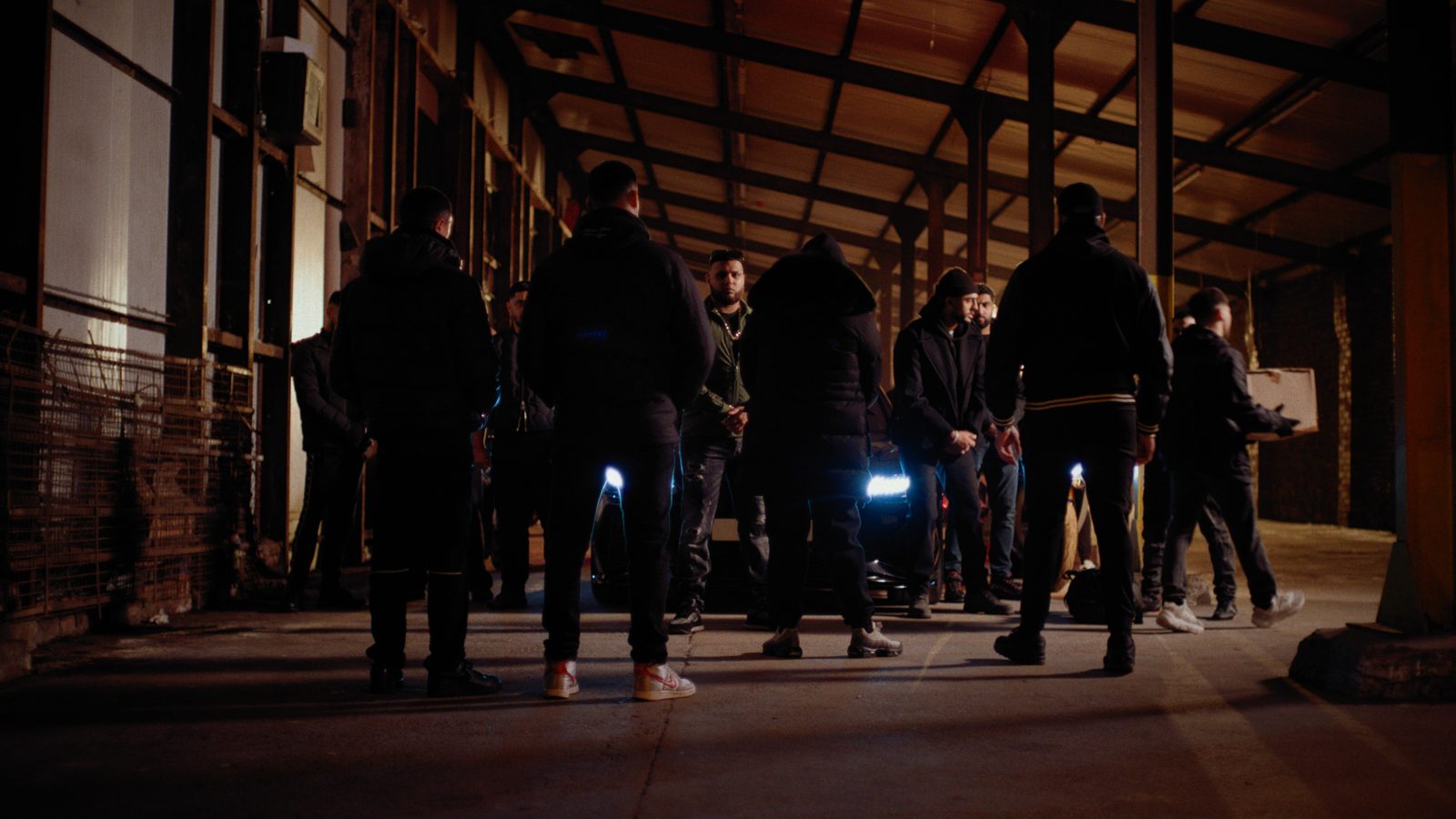 A still from Apnas: a group of men gathered around a car in a dark warehouse at night